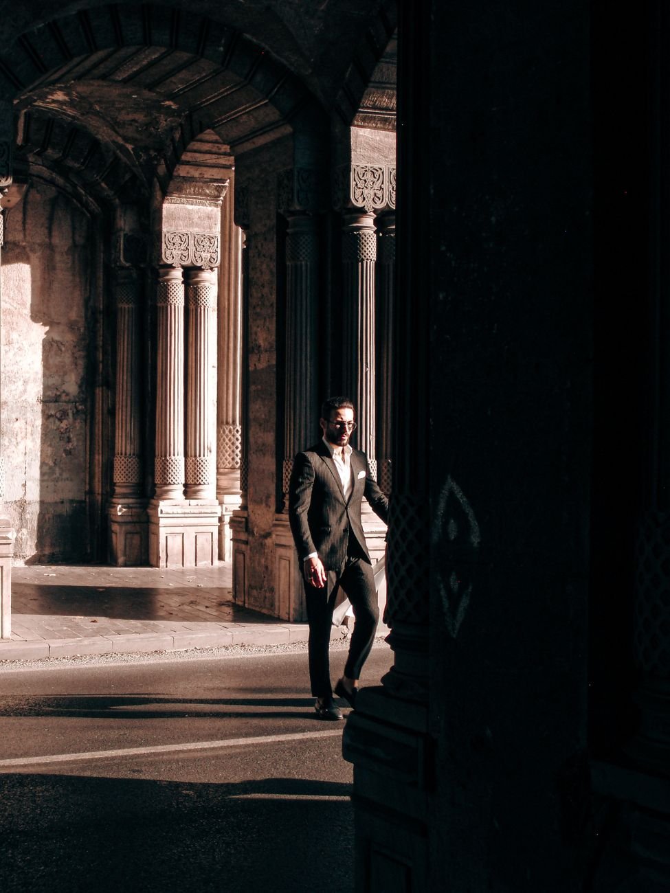 Elegant man in a suit walks through a historic archway cast in dramatic shadows. Urban exploration.